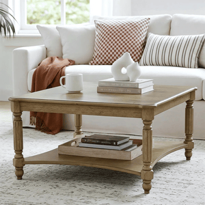 A wooden square coffee table with turned legs features a shelf below. It is styled with books, a white abstract sculpture, and a mug. The background includes a beige couch with checkered and striped pillows and a brown throw blanket.