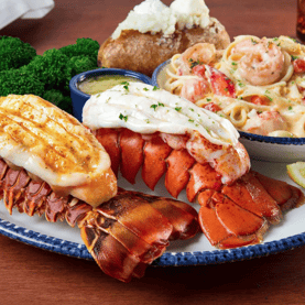 Grilled and steamed lobster tails with lemon wedges, creamy shrimp pasta, a baked potato with butter, broccoli, and a cup of sauce are arranged on a blue-edged plate.