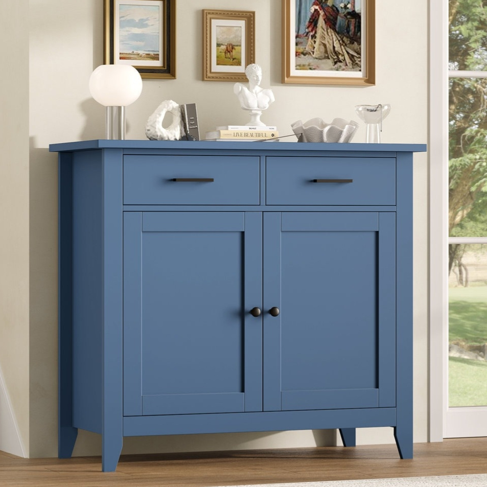 A blue sideboard with two drawers and cabinet doors is set against a wall with framed artwork. It holds decorative items, including a sculpture and a lamp, and is positioned near a window and staircase.