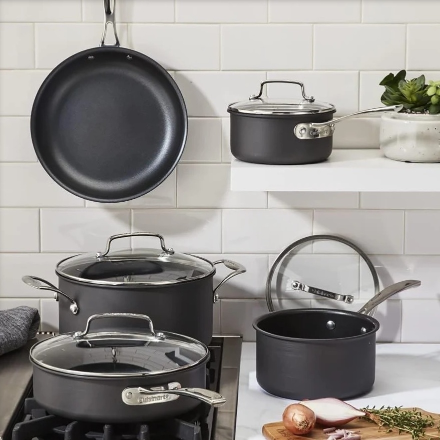 Cookware set with frying pan, saucepans, and stock pot, all with glass lids and metal handles, displayed on a stove and shelf with a white tiled backsplash and some kitchen herbs and an onion on a cutting board nearby.