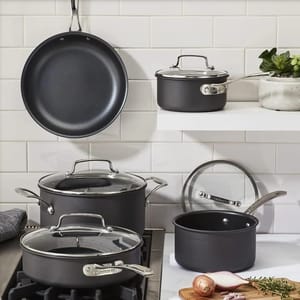 Cookware set with frying pan, saucepans, and stock pot, all with glass lids and metal handles, displayed on a stove and shelf with a white tiled backsplash and some kitchen herbs and an onion on a cutting board nearby.