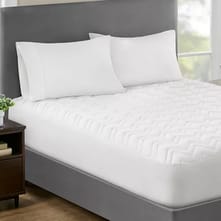 A neatly made bed features a deep-pocket mattress pad with a white, textured, quilted design. The pad is fitted on a mattress with a gray upholstered headboard, accompanied by two white pillows, and a wooden nightstand beside it.