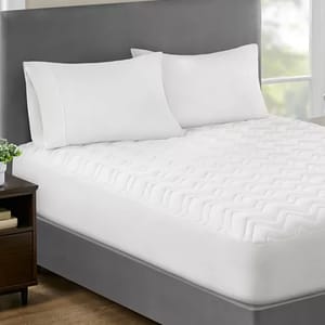 A neatly made bed features a deep-pocket mattress pad with a white, textured, quilted design. The pad is fitted on a mattress with a gray upholstered headboard, accompanied by two white pillows, and a wooden nightstand beside it.