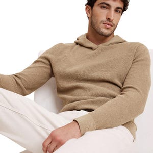 A man wears a beige, textured knit hoodie paired with white pants, featuring a relaxed and casual design.
