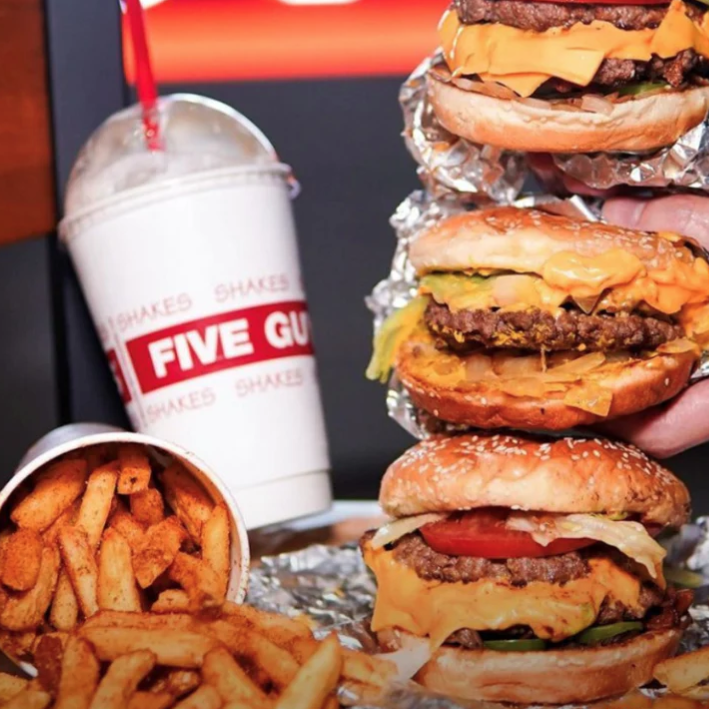 Burgers with cheese and toppings, a cup of seasoned fries, and a milkshake with a red straw from Five Guys, with visible branding on the cup.