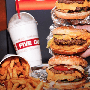 Burgers with cheese and toppings, a cup of seasoned fries, and a milkshake with a red straw from Five Guys, with visible branding on the cup.