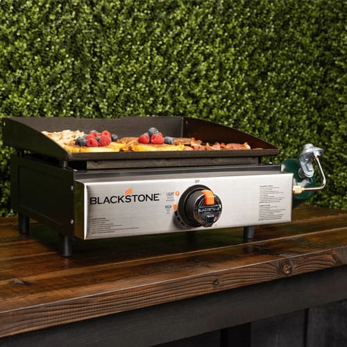 Blackstone outdoor griddle with food on cooking surface, positioned on a wooden table against a leafy backdrop.