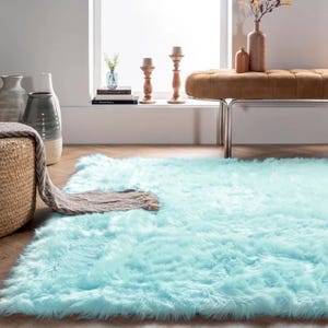 A light blue fluffy rug adorns a wood floor, accompanied by a wicker basket, a soft gray blanket, decorative vases, and a brown bench near a bright window.