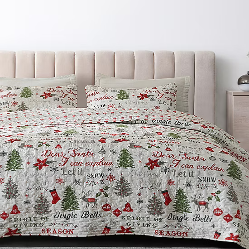 A holiday-themed quilt with matching pillow shams featuring festive phrases and designs, such as Christmas trees, poinsettias, and ornaments, in red, green, and white.