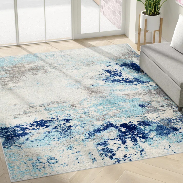A modern area rug with an abstract design features shades of blue, gray, and white on a floor next to a sofa and plant.