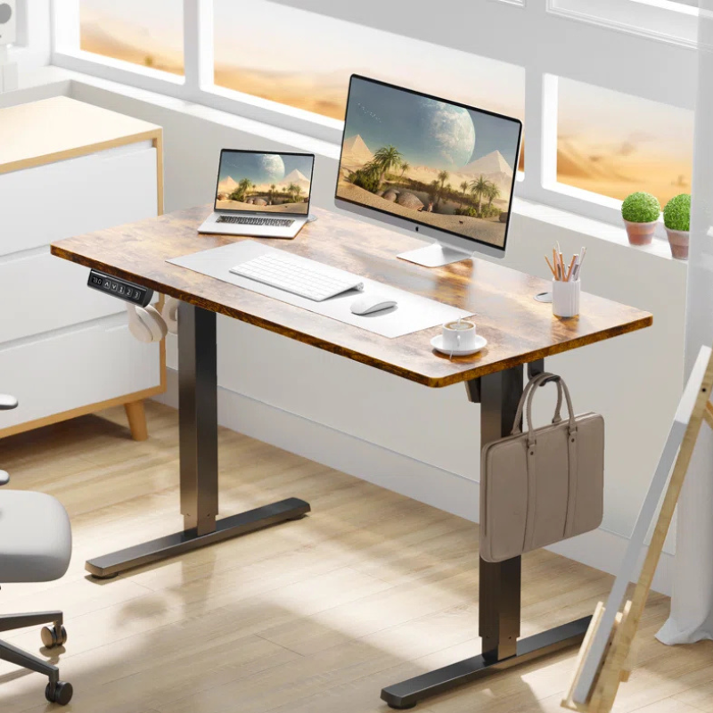 A modern adjustable standing desk with a wood finish features a laptop, monitor, keyboard, mouse, and cup of coffee. A bag hangs on the desk side, and a headset rests on the frame. Potted plants and a pencil holder sit near the desk.