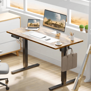 A modern adjustable standing desk with a wood finish features a laptop, monitor, keyboard, mouse, and cup of coffee. A bag hangs on the desk side, and a headset rests on the frame. Potted plants and a pencil holder sit near the desk.