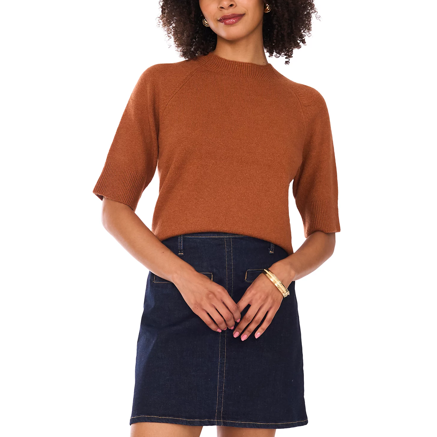 A woman wearing a short-sleeve, rust-colored sweater paired with a dark blue denim skirt. She accessorizes with gold hoop earrings and matching bangles.