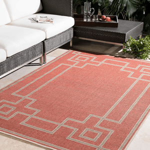 Red geometric-patterned area rug placed in a modern outdoor setting, partially under a wicker sofa with white cushions and next to a black coffee table holding a glass jug and apples.