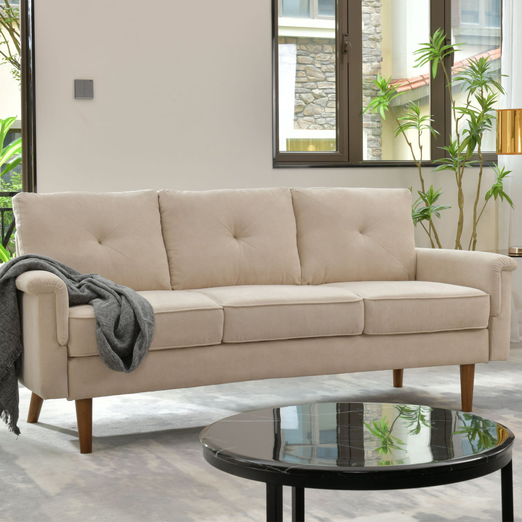 A beige 72\“ upholstered sofa with a tufted backrest and three seat cushions, complemented by wooden legs and a gray throw draped over one arm. It is situated in a modern living space with a round glass coffee table and indoor plants.