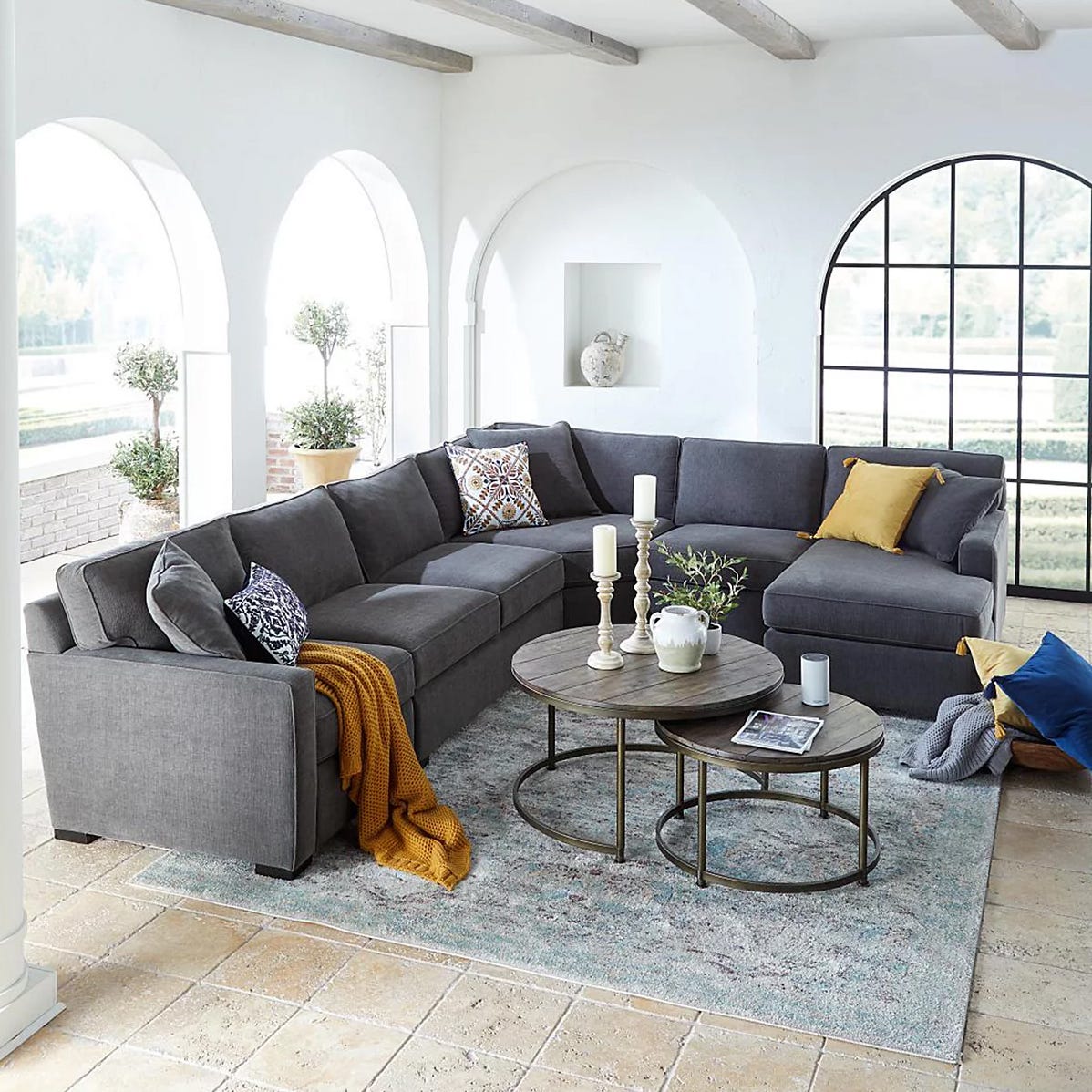 A gray sectional sofa with colorful pillows sits in an airy room featuring arched windows, decorative candles on round coffee tables, a mustard throw, and a textured rug.
