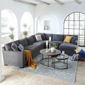 A gray sectional sofa with colorful pillows sits in an airy room featuring arched windows, decorative candles on round coffee tables, a mustard throw, and a textured rug.