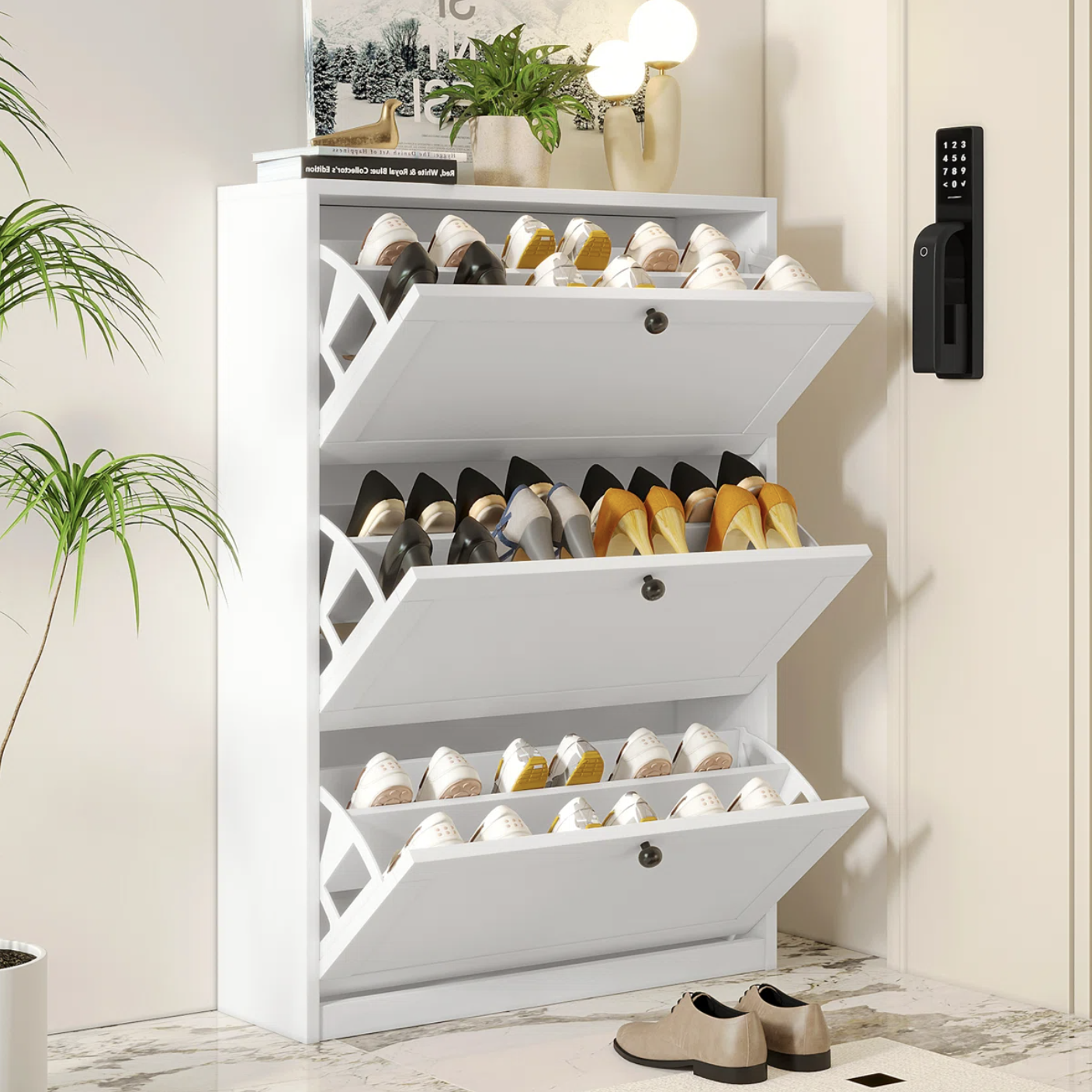 A white, multi-tiered shoe cabinet with drop-down compartments holds various shoes. It is placed next to a keypad door lock and is styled with a lamp, plant, and decorative items on top.