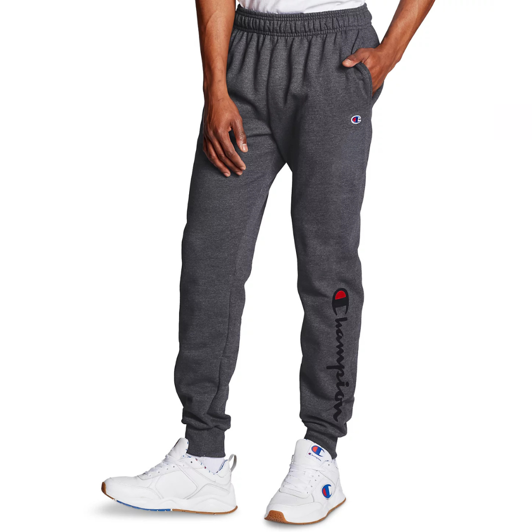 Dark gray jogger pants with an elastic waistband and cuffs, featuring the Champion logo embroidered on the thigh and printed along the lower leg. Worn with white sneakers also displaying the Champion logo.