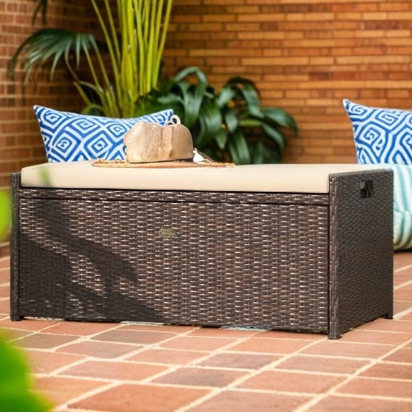 Outdoor rattan storage bench with a beige cushion, featuring a dark woven design, set on a brick patio with plants and blue patterned pillows.