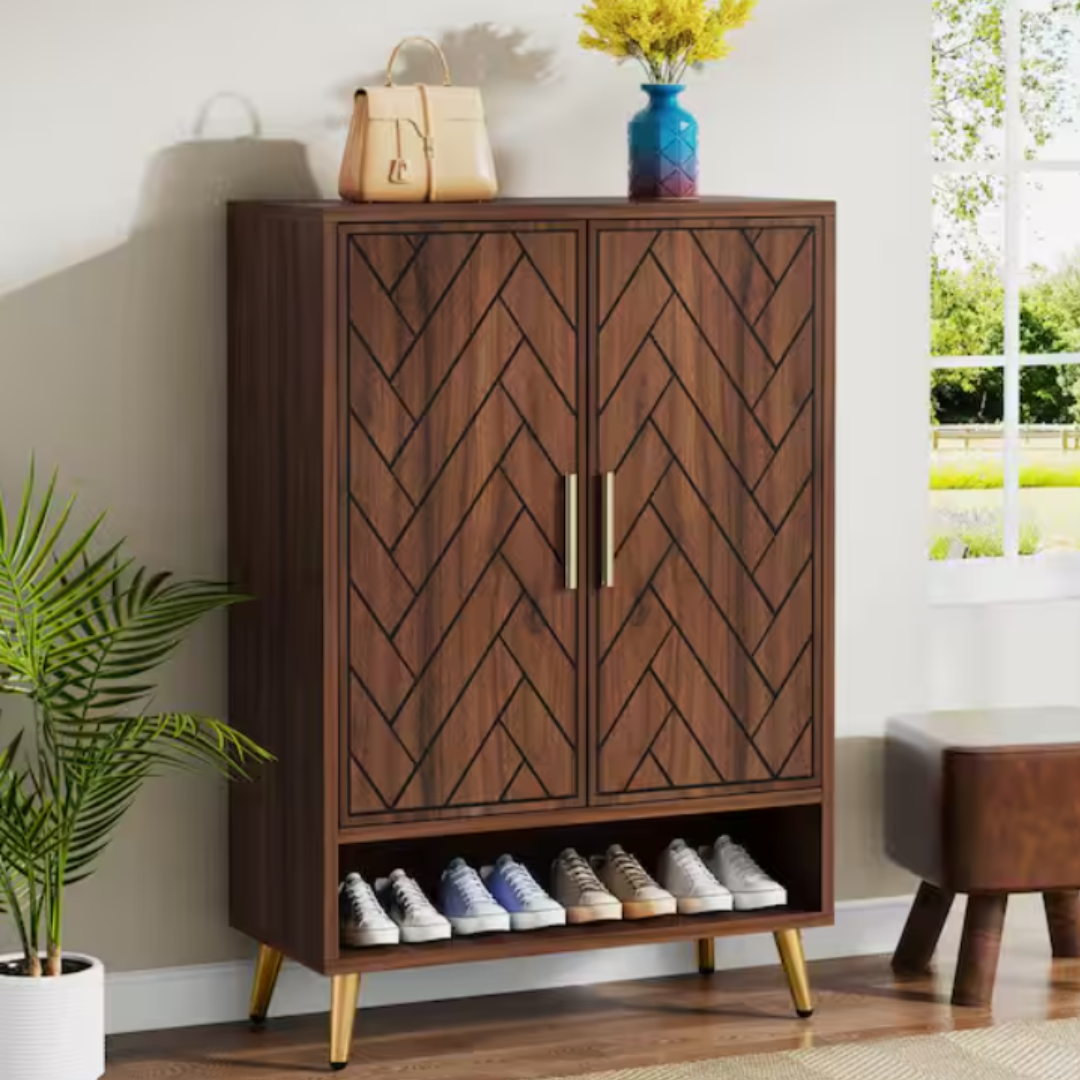 A wooden cabinet with a chevron pattern features gold handles and legs. It includes a bottom shelf for storing shoes and is topped with a handbag and a vase with yellow flowers.