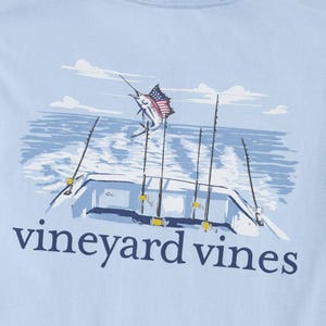 Light blue Vineyard Vines shirt featuring a graphic of a fishing boat with rods and a leaping marlin with an American flag pattern on its fin.