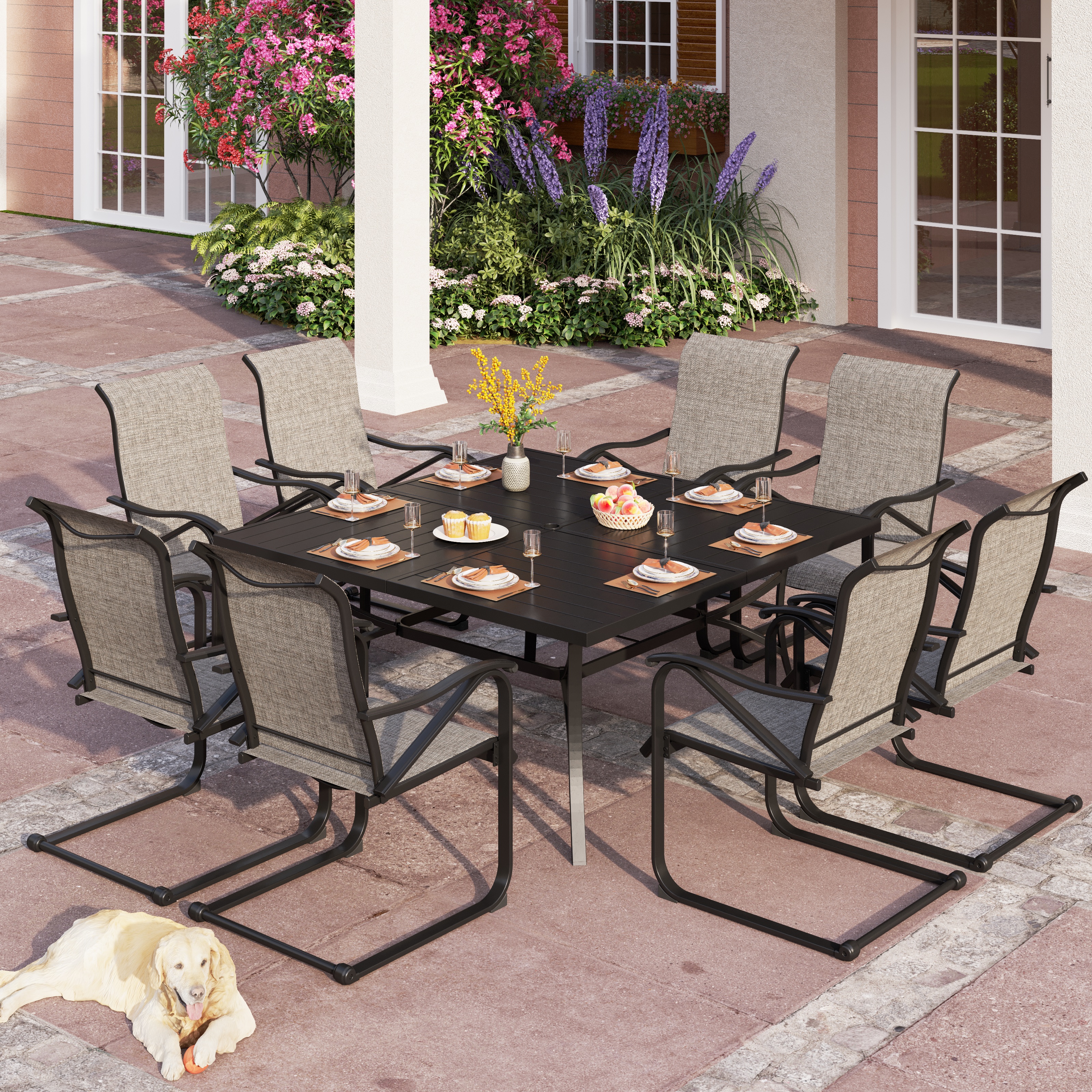 A 9-piece patio dining set featuring a square black table and eight gray cushioned chairs with black frames. The table is set with plates, glasses, and a vase with yellow flowers, and a golden retriever lies nearby on a tiled patio.