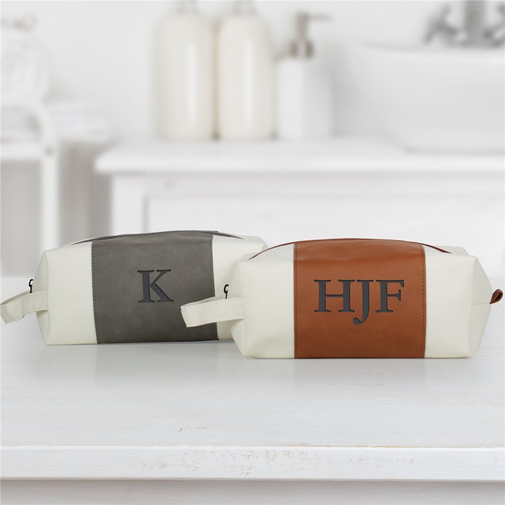 Two toiletry bags, one with a gray stripe and single initial \“K,\“ and the other with a brown stripe and initials \“HJF,\“ are displayed on a white countertop. Both have handles and zippers.