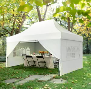 White outdoor canopy tent with side walls and windows, set up over a table with chairs in a garden setting.