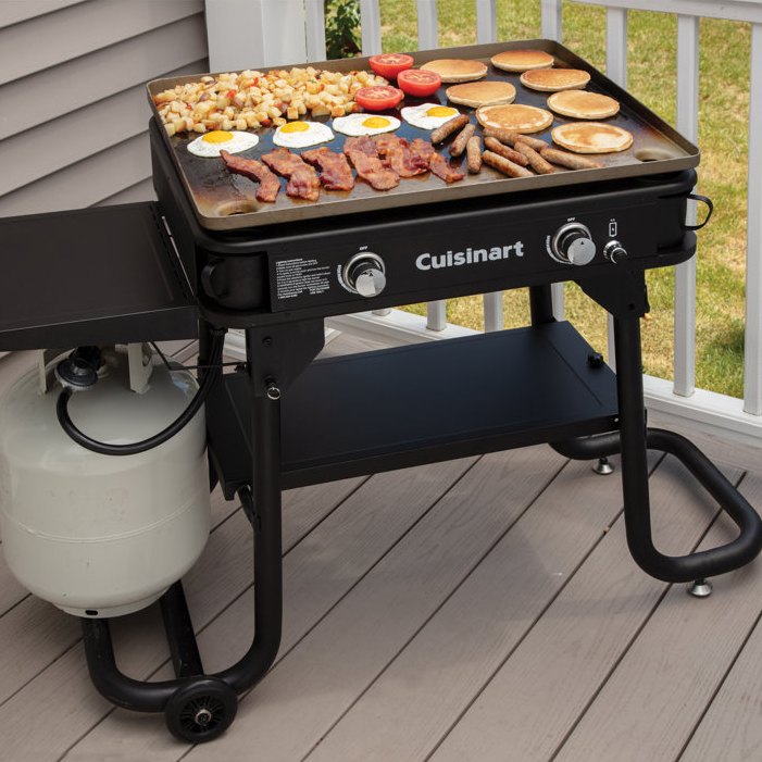 Cuisinart 28\“ Gas Griddle with Cover features a large cooking surface with two burners, ideal for grilling various breakfast items like bacon, eggs, pancakes, and sausages. It includes a propane tank connection and wheels for mobility.