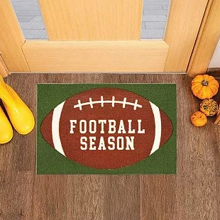 A doormat featuring a football design with \“FOOTBALL SEASON\“ text, placed by a door with pumpkins and yellow boots nearby.