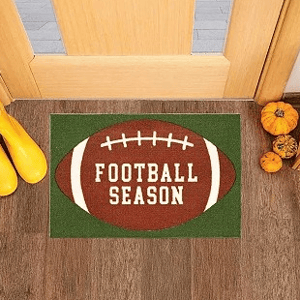 A doormat featuring a football design with \“FOOTBALL SEASON\“ text, placed by a door with pumpkins and yellow boots nearby.