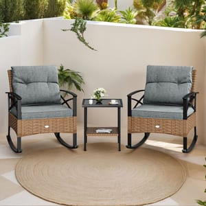 Two wicker rocking chairs with gray cushions sit beside a small black table, featuring a plant and cups, on a patio area with plants and a circular rug.