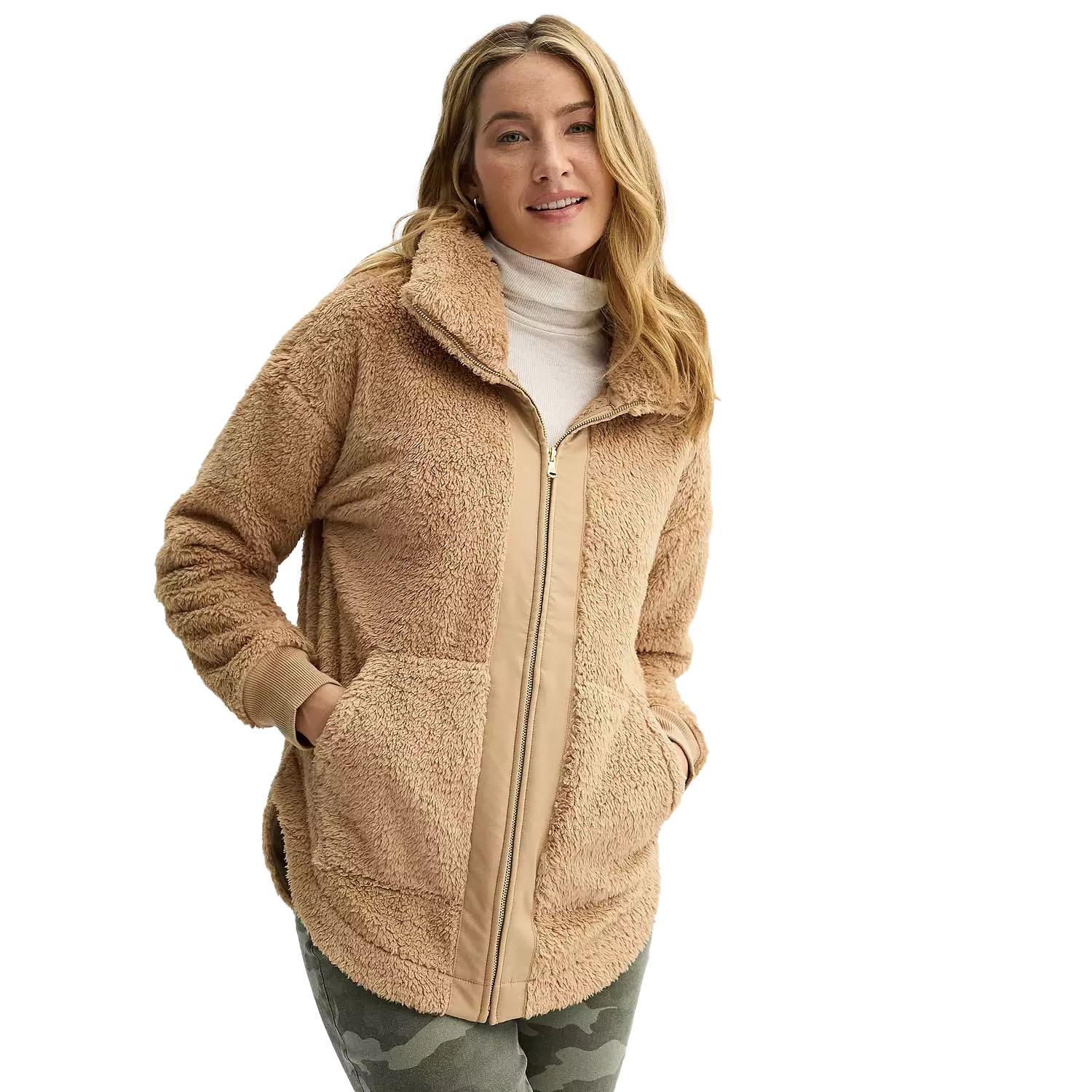 A woman models a beige sherpa jacket with a zippered front, side pockets, and a raised collar, paired with a white turtleneck and camo pants.