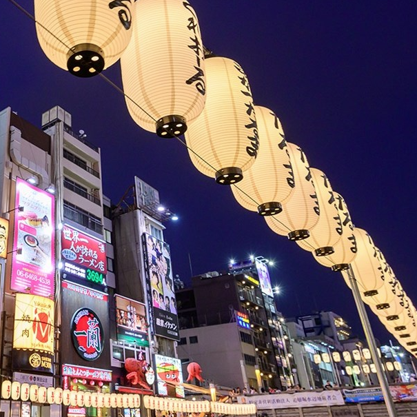 A nighttime street scene with illuminated lanterns hanging over buildings in a lively area, featuring colorful signs for restaurants and shops.