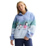 A woman is wearing a cozy, light blue fleece pullover with a playful, winter-themed design featuring children. The pullover has a zippered collar and is paired with blue pants.