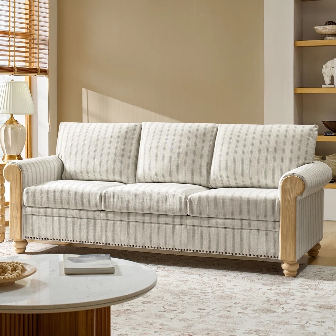 A striped sofa with wooden accents and rounded arms is placed in a living room setting, complemented by a nearby lamp and a round marble-topped coffee table.