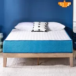 A bed with a blue mattress and white top sits on a wooden frame. It is accompanied by white pillows and a patterned black-and-white cushion, with a blue wall background and an orange hanging light.