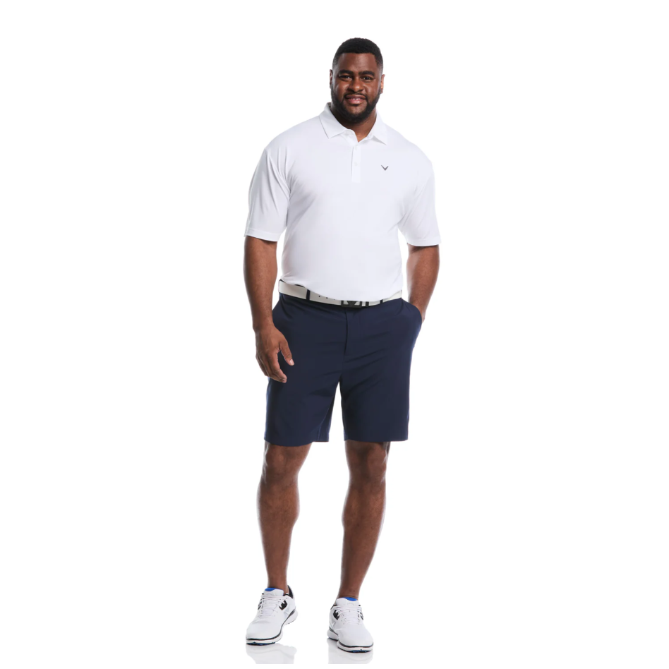A man is wearing a white polo shirt, navy shorts, a black belt, and white sneakers.