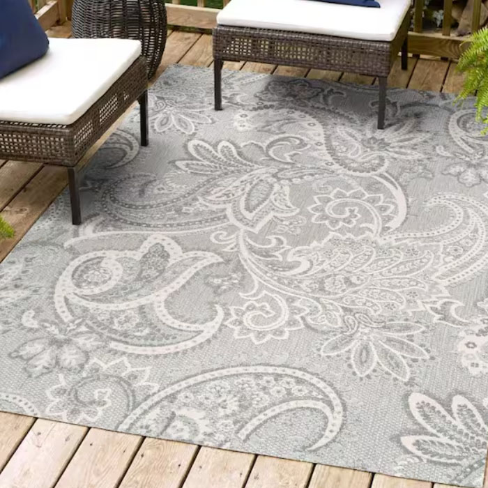 A grey outdoor rug with a paisley pattern is placed on a wooden deck. It's surrounded by wicker chairs with white cushions and blue accent pillows.