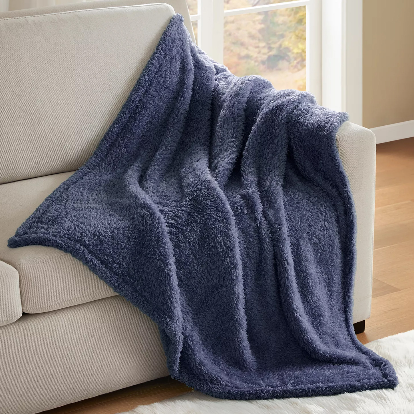 A plush, dark blue sherpa throw blanket is draped over a light-colored sofa in a room with a window overlooking an outdoor scene.