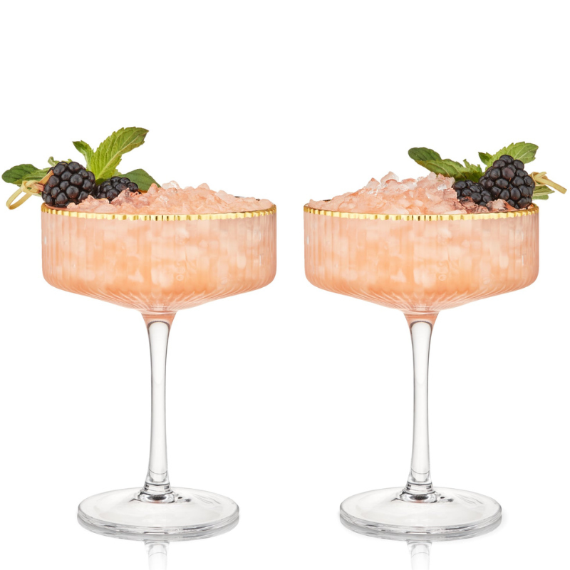 Viski Meridian Crystal Coupe Set features two elegant coupes with a ribbed texture and gold rim, each filled with a pink beverage topped with crushed ice. Garnished with blackberries and mint leaves on the side for added presentation.