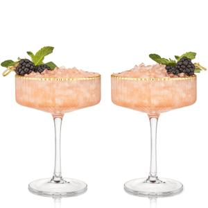 Viski Meridian Crystal Coupe Set features two elegant coupes with a ribbed texture and gold rim, each filled with a pink beverage topped with crushed ice. Garnished with blackberries and mint leaves on the side for added presentation.