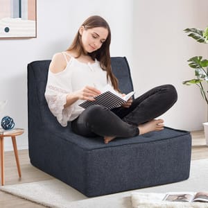 A woman sits on a dark blue floor chair while reading a book in a cozy room with a small table, decorative objects, and a plant.