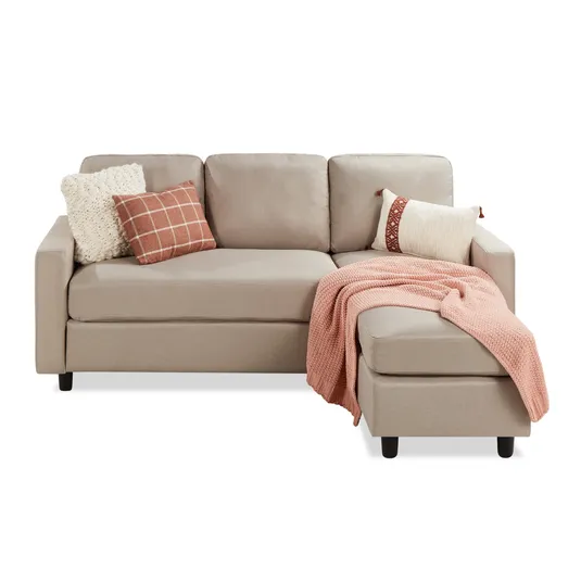 Beige L-shaped sofa with three cushion backs, adorned with a cream and a checkered brown pillow. A pink knit throw blanket is draped over the chaise section.