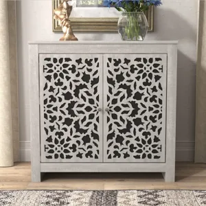 A two-door cabinet with an intricate cutout pattern on the doors, featuring a light wood frame. It is topped with a decorative sculpture and a glass vase with blue flowers.