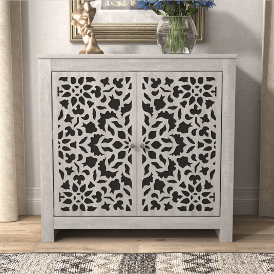 A two-door cabinet with an intricate cutout pattern on the doors, featuring a light wood frame. It is topped with a decorative sculpture and a glass vase with blue flowers.