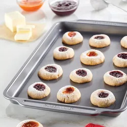 A nonstick baking sheet displays thumbprint cookies filled with apricot and raspberry jam, accompanied by butter and jam bowls in the background.