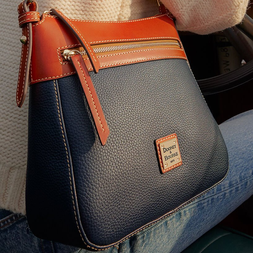 The Dooney & Bourke Pebble Crossbody features a textured navy body with contrast tan leather trim, top zipper, and outer pocket. A gold metal logo plate is on the front, highlighted by white stitching.