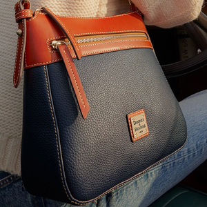 The Dooney & Bourke Pebble Crossbody features a textured navy body with contrast tan leather trim, top zipper, and outer pocket. A gold metal logo plate is on the front, highlighted by white stitching.
